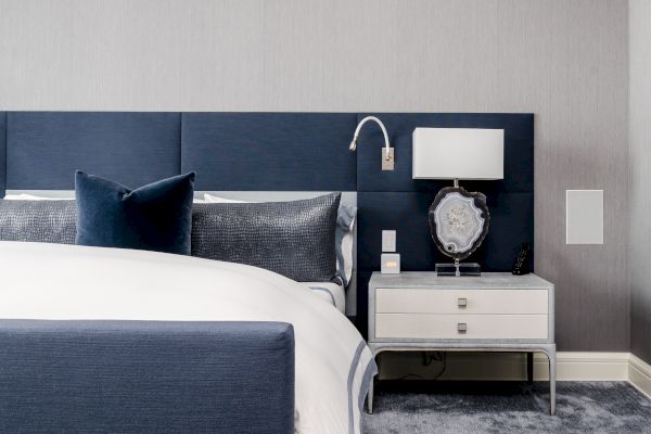 A modern bedroom with a navy blue upholstered bed, a white and blue nightstand holding a lamp and photo frame, and a gray carpet.