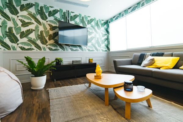 A modern living room with a tropical leaf wallpaper, a wall-mounted TV, a gray sofa with yellow cushions, wooden tables, and indoor plants.