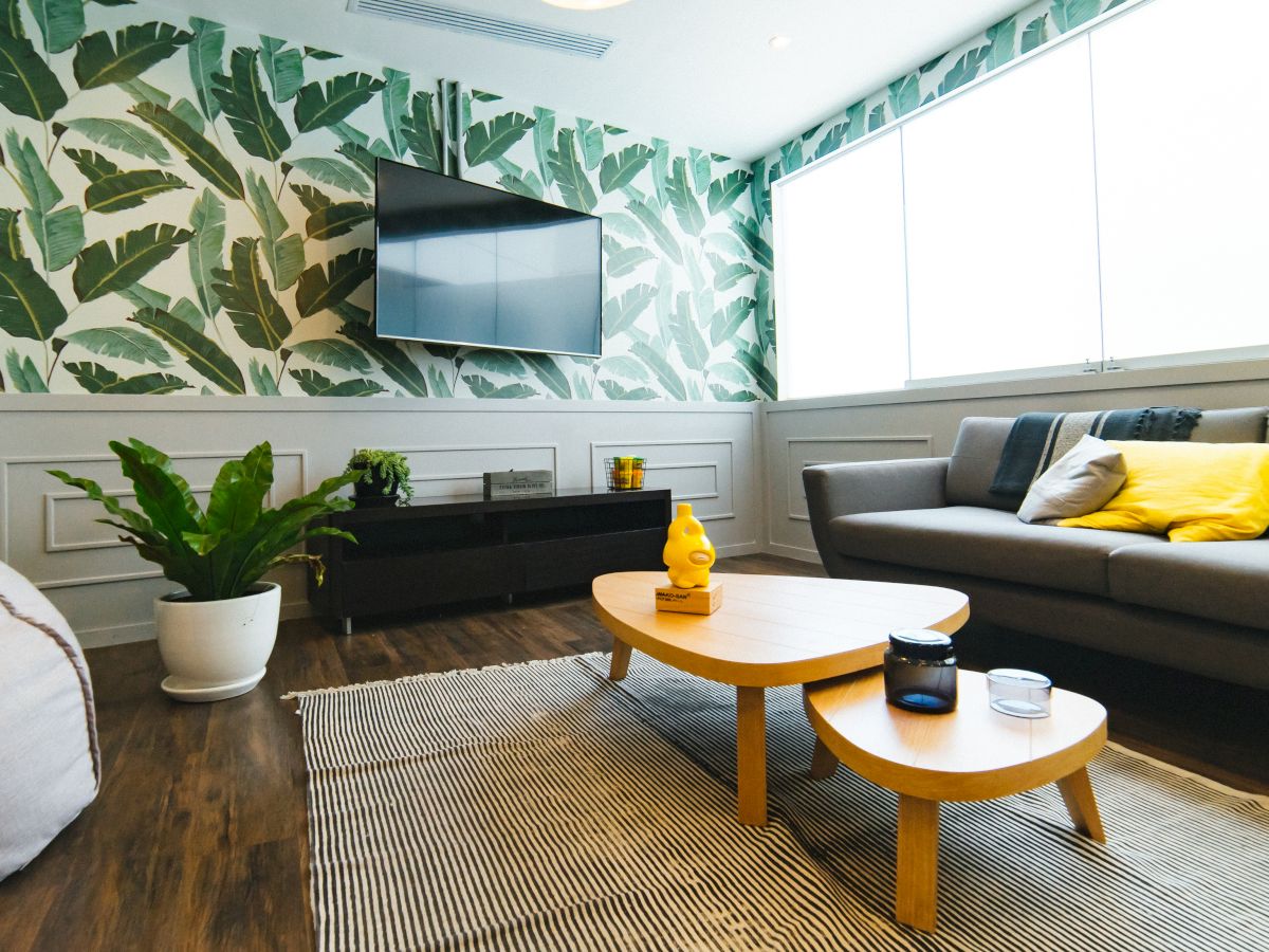 A modern living room with a gray sofa, yellow cushions, a TV, a rug, a potted plant, and a coffee table, featuring leaf-patterned wallpaper.