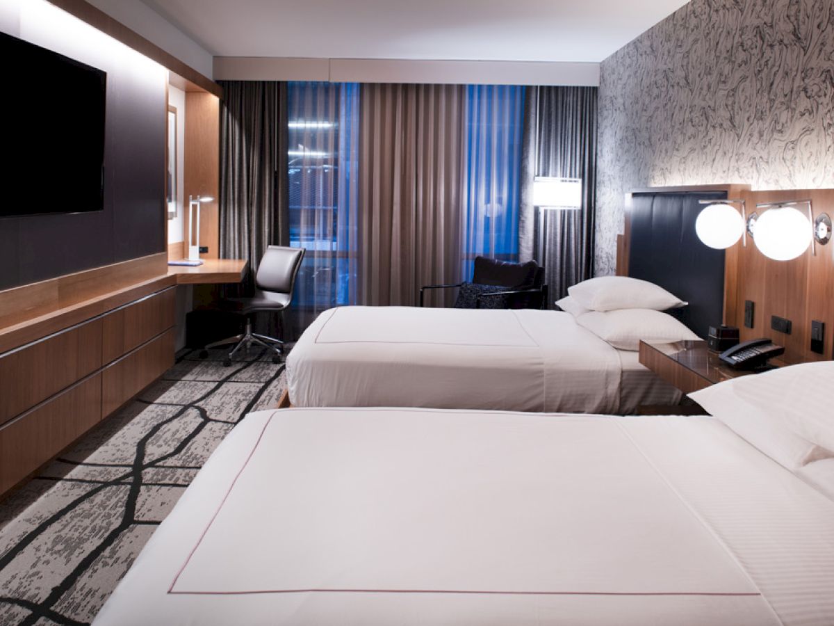 The image shows a modern hotel room featuring two double beds, a wall-mounted TV, a work desk with a chair, and contemporary decor ending the sentence.
