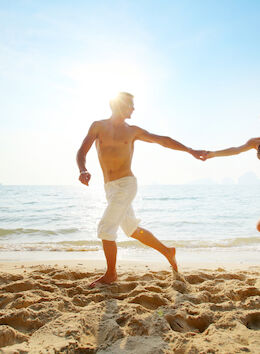 A man and a woman are running and holding hands on a sandy beach with the ocean and sunlight in the background.