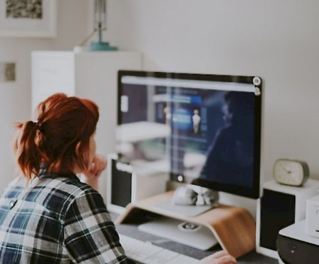 A person with red hair sits at a desk, looking at a computer screen. The desk has a lamp, speakers, a clock, and some storage items.