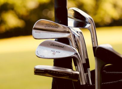 The image shows a close-up of a set of golf clubs in a golf bag with lush green grass and trees in the background.