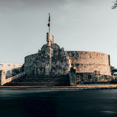 A stone monument with steps leading up to it, topped with a flagpole flying a flag. The structure is circular with intricate carvings.