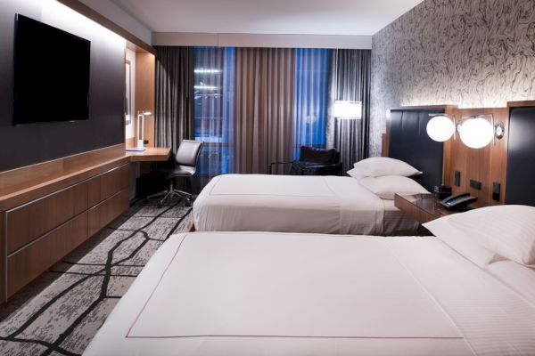 The image shows a modern hotel room with two beds, a wall-mounted TV, a desk with a chair, and contemporary lighting and decor on one wall.