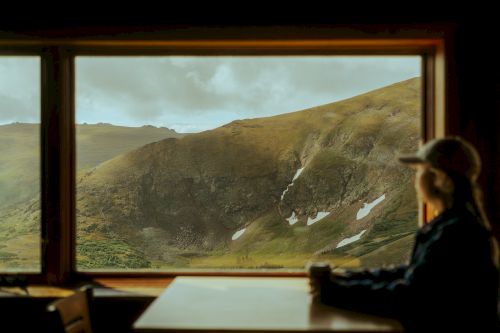 A person sits at a table inside, looking out through a large window at a scenic mountainous landscape with patches of snow.