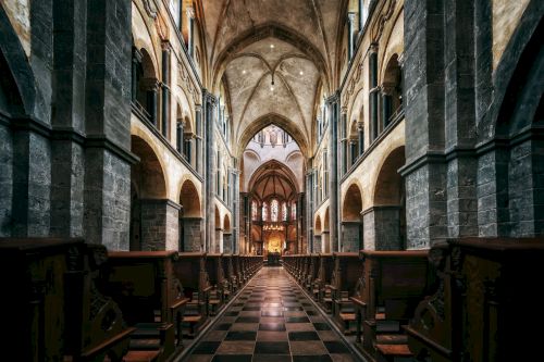 A beautifully lit Gothic church interior with vaulted ceilings, arches, pillars, and symmetrical rows of wooden pews along a central aisle ending the sentence.