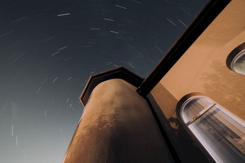 The image shows a night sky with star trails captured above a tall building with arched windows, set against a dark background.