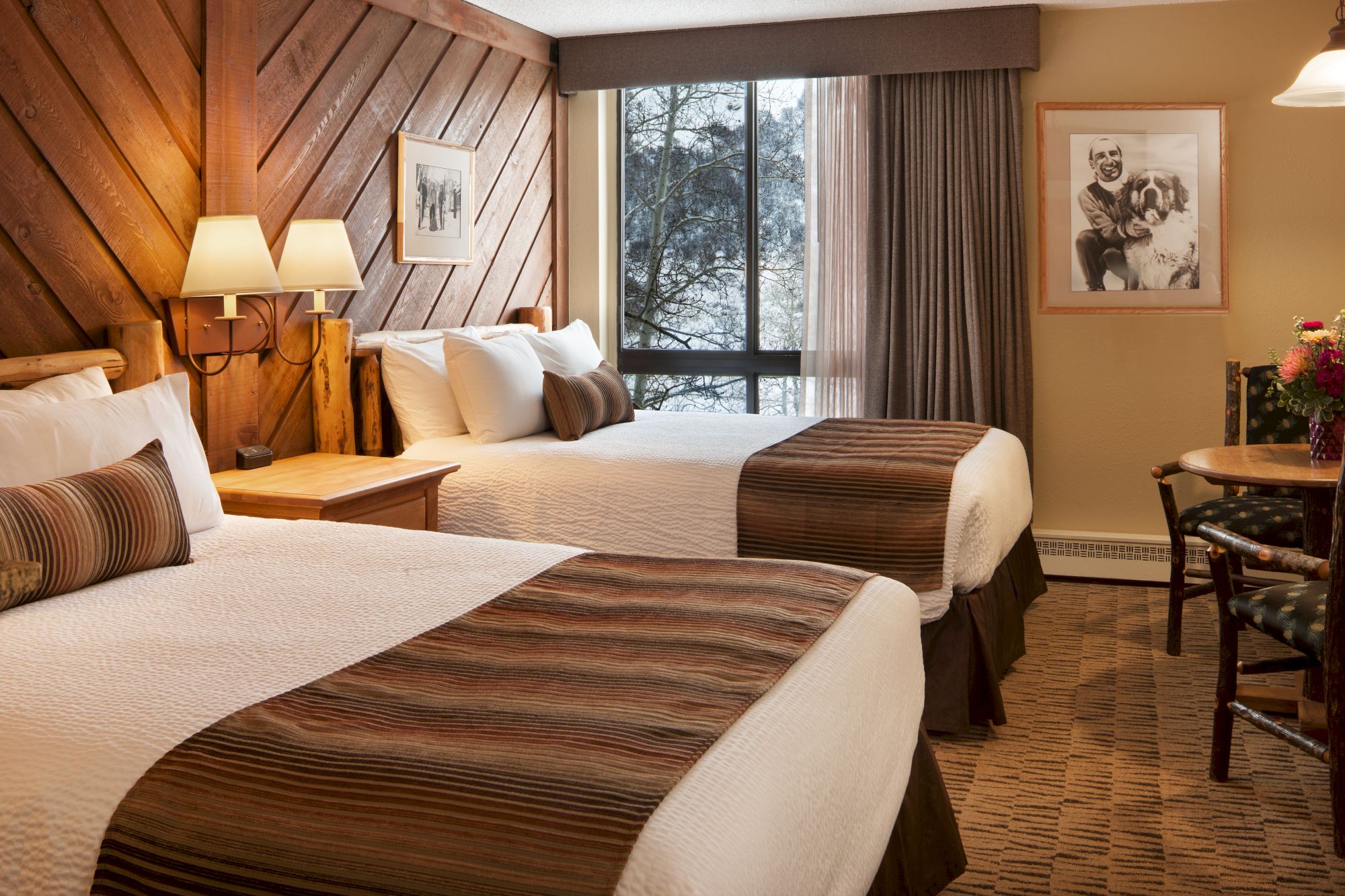 The image shows a cozy hotel room featuring two double beds with brown accents, a wood-paneled wall, a window with a view, and a small seating area.