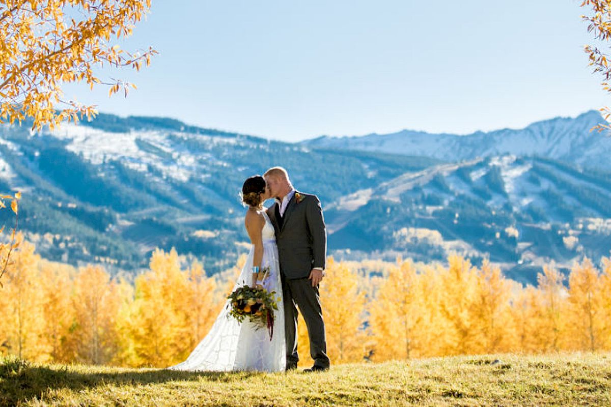 A couple kisses in a scenic outdoor setting with mountains in the background and trees displaying autumn colors.