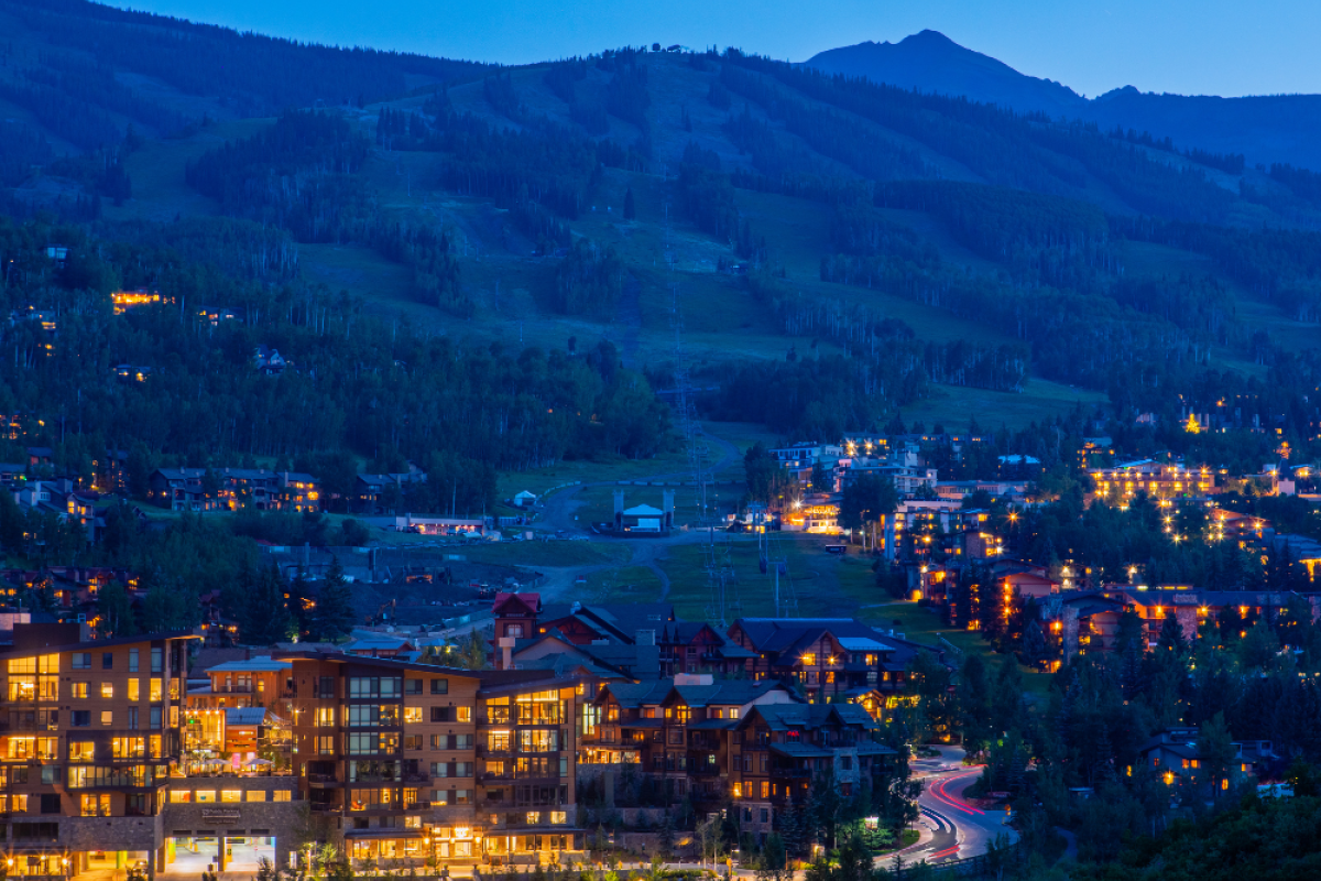 The image shows a scenic evening view of a town nestled in the mountains, with illuminated buildings and houses, surrounded by lush greenery and hills.
