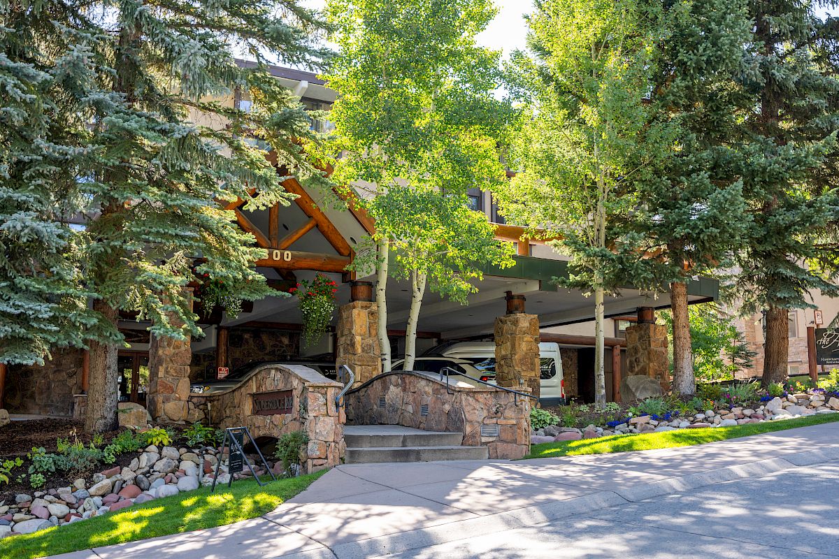 A building entrance surrounded by trees, with stone pillars and a stone wall. A sign for "The Pines Lodge" is visible on the right side of the image.