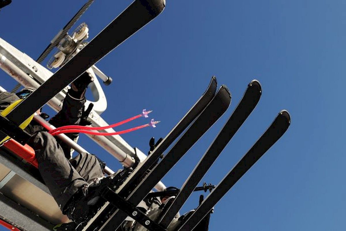 A close-up of ski equipment on a ski lift against a clear blue sky, with trees visible in the corner.