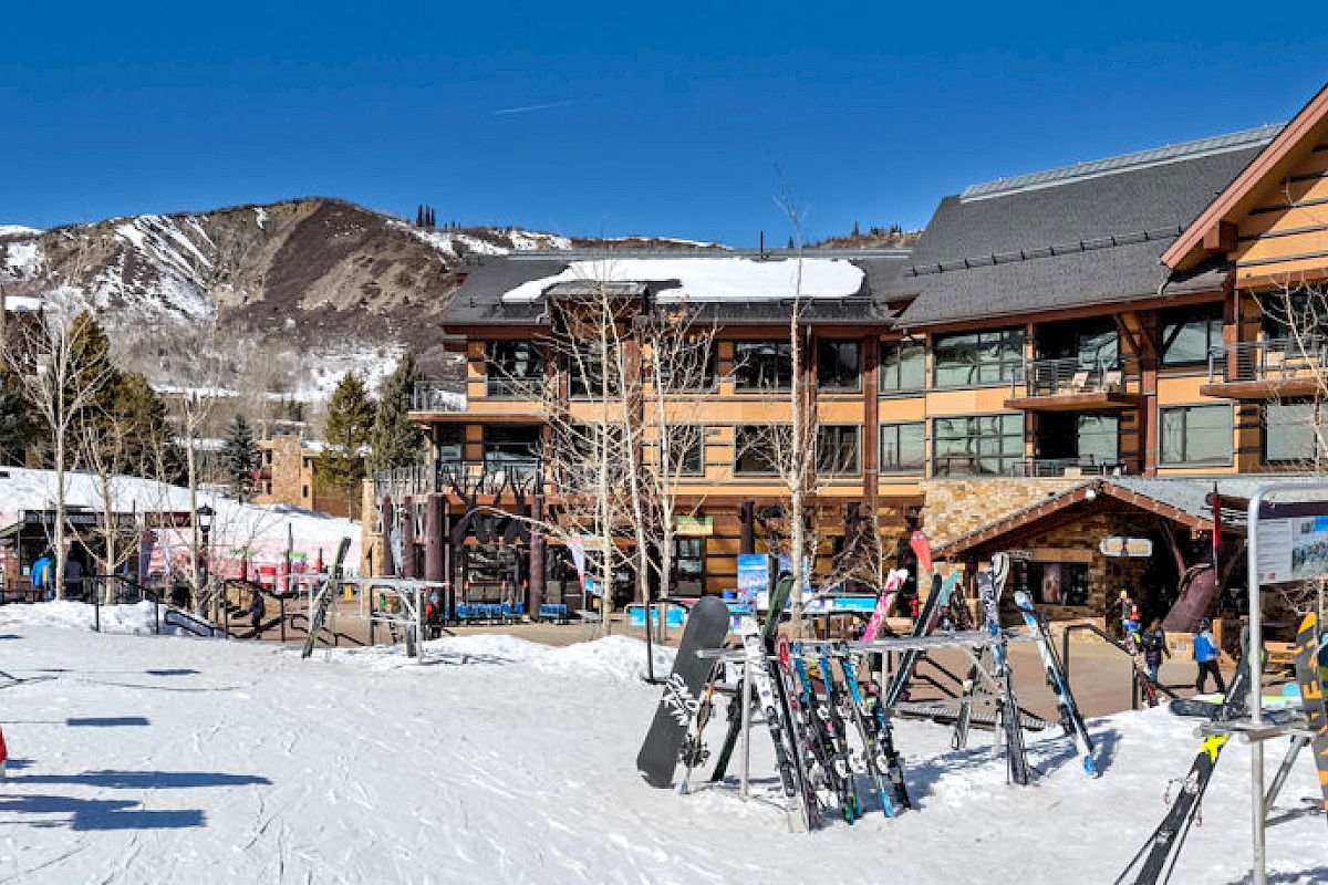 This image shows a snowy ski resort with skiers and snowboarders getting ready near a ski school and lodge. Snow-covered mountains are in the background.
