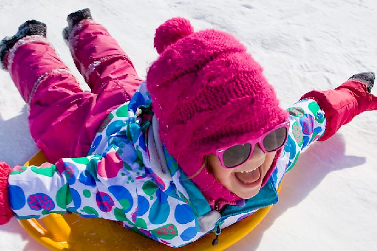 A child dressed in colorful winter clothing and pink hat gleefully sleds down a snowy hill on a yellow sled while smiling widely.