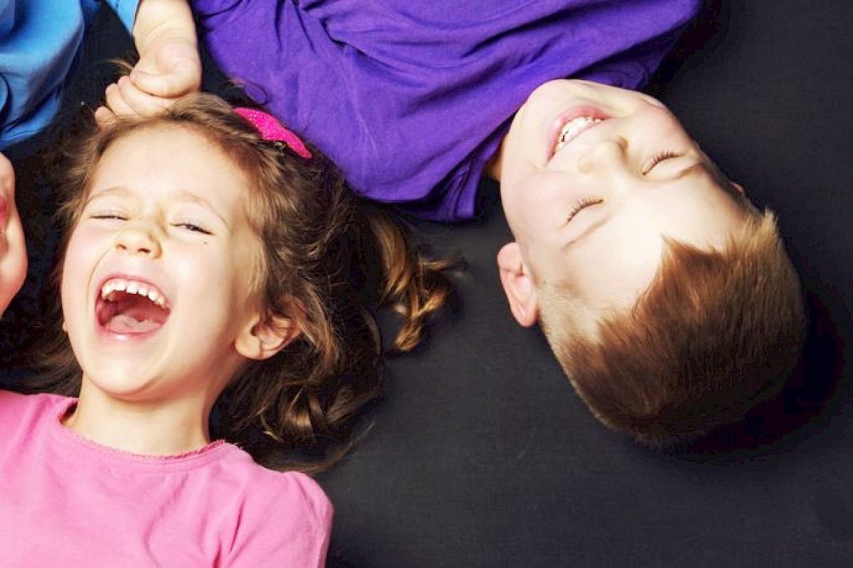 Three children lie on their backs, laughing joyfully while holding hands. The background appears to be a dark, plain surface.