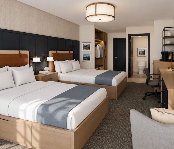 Two queen beds in a modern hotel room, with a desk, TV, armchair, neutral tones, ample lighting, and a small hallway leading to a door.