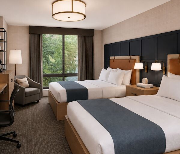 Two double beds with white linens and navy runners in a modern hotel room, plus a desk, chair, flat screen, and a balcony view.