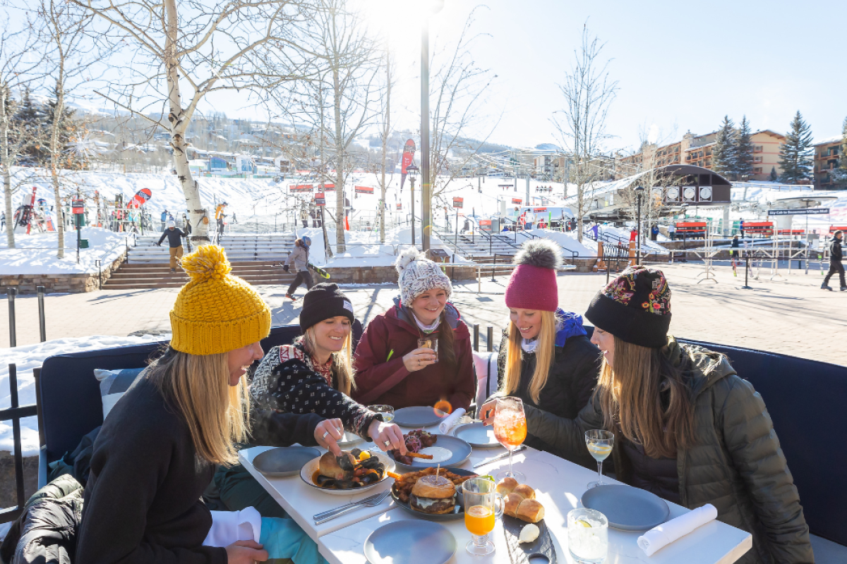 A group of people enjoying a meal outdoors in a snowy area, all dressed warmly in winter attire, with a bright sun shining above them.