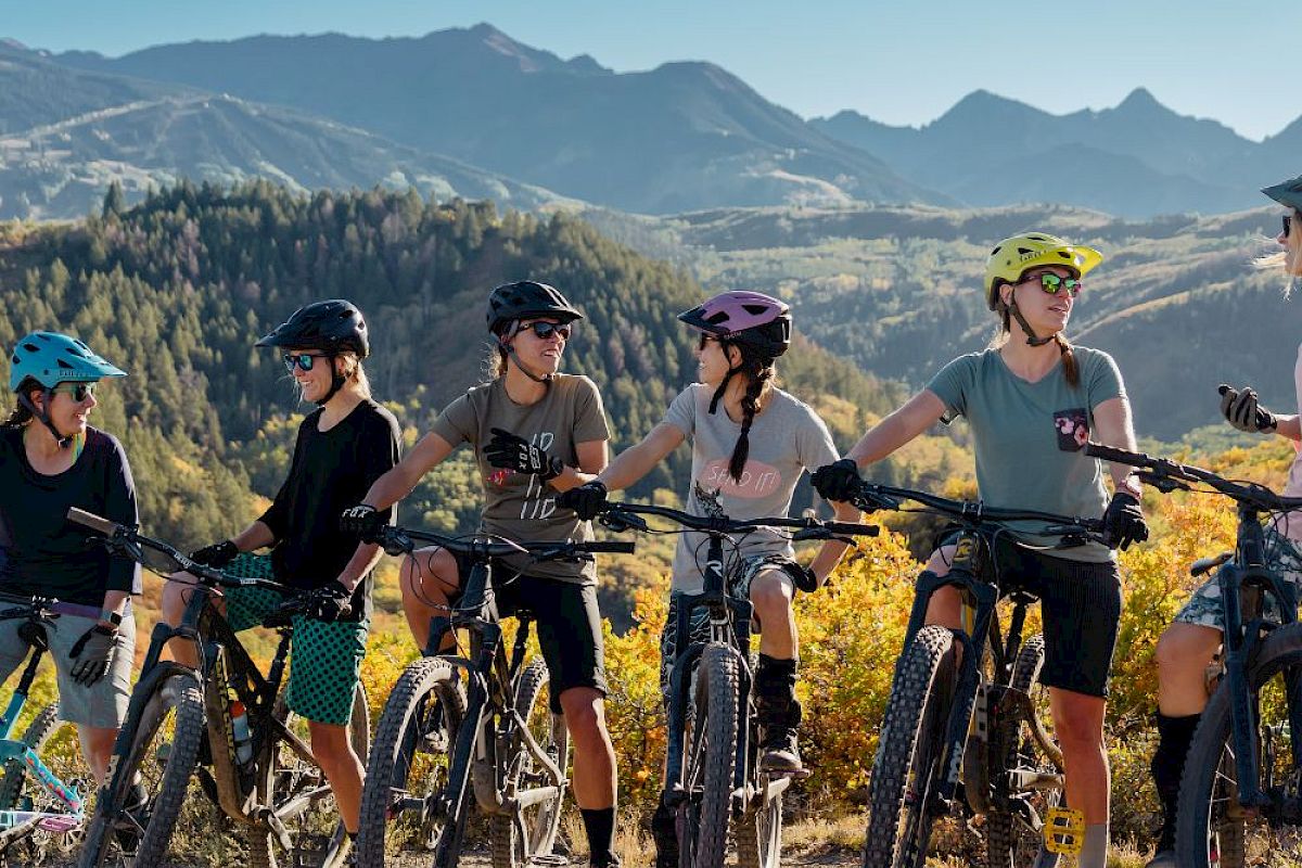A group of cyclists, wearing helmets, stands with their bikes on a scenic mountain trail, engaging in conversation with a beautiful view in the background.