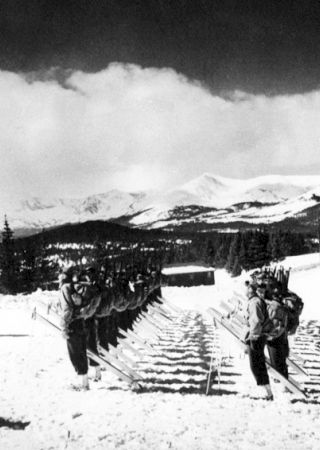 The image shows a group of soldiers in winter gear, skiing in a snowy mountainous landscape. The background features snow-covered peaks and evergreen trees.