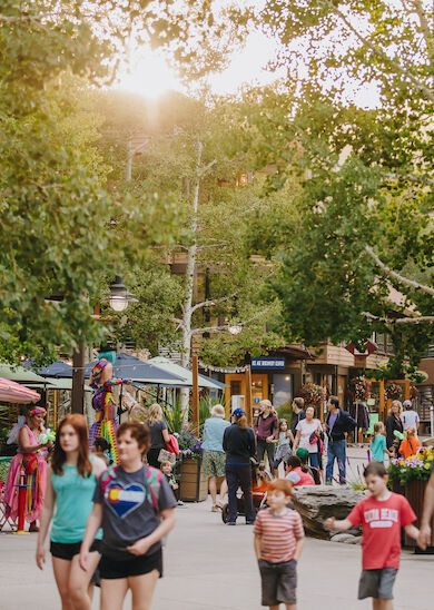 People are walking on a tree-lined village street with shops and outdoor seating, enjoying a sunny day.