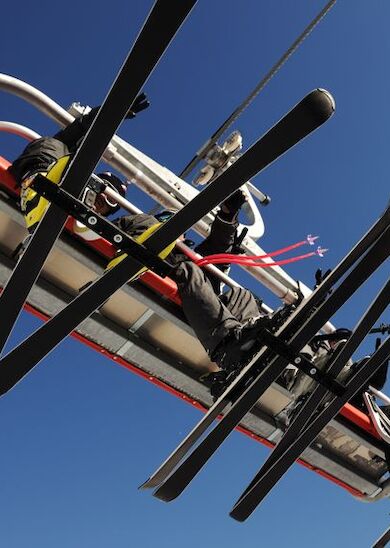 Two people sitting on a ski lift with skis attached, viewed from below against a clear blue sky.