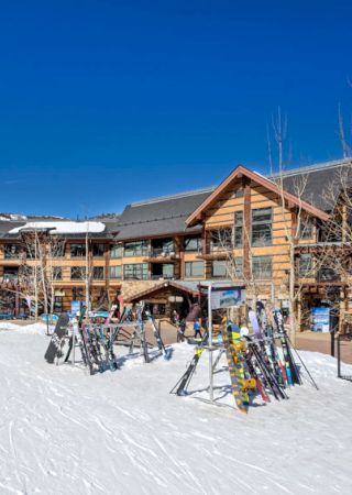 The image shows a snowy ski resort with a wooden lodge, ski racks filled with skis, and a clear blue sky in the background.