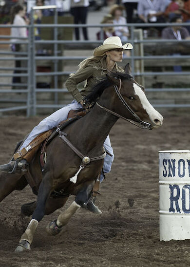 The image shows a rider on a horse making a sharp turn around a barrel labeled "Snowman Rodeo" in a rodeo arena.