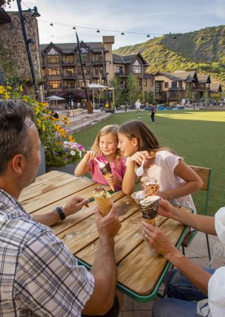 A family is seated at an outdoor table enjoying ice cream cones with a scenic backdrop of buildings and mountains.