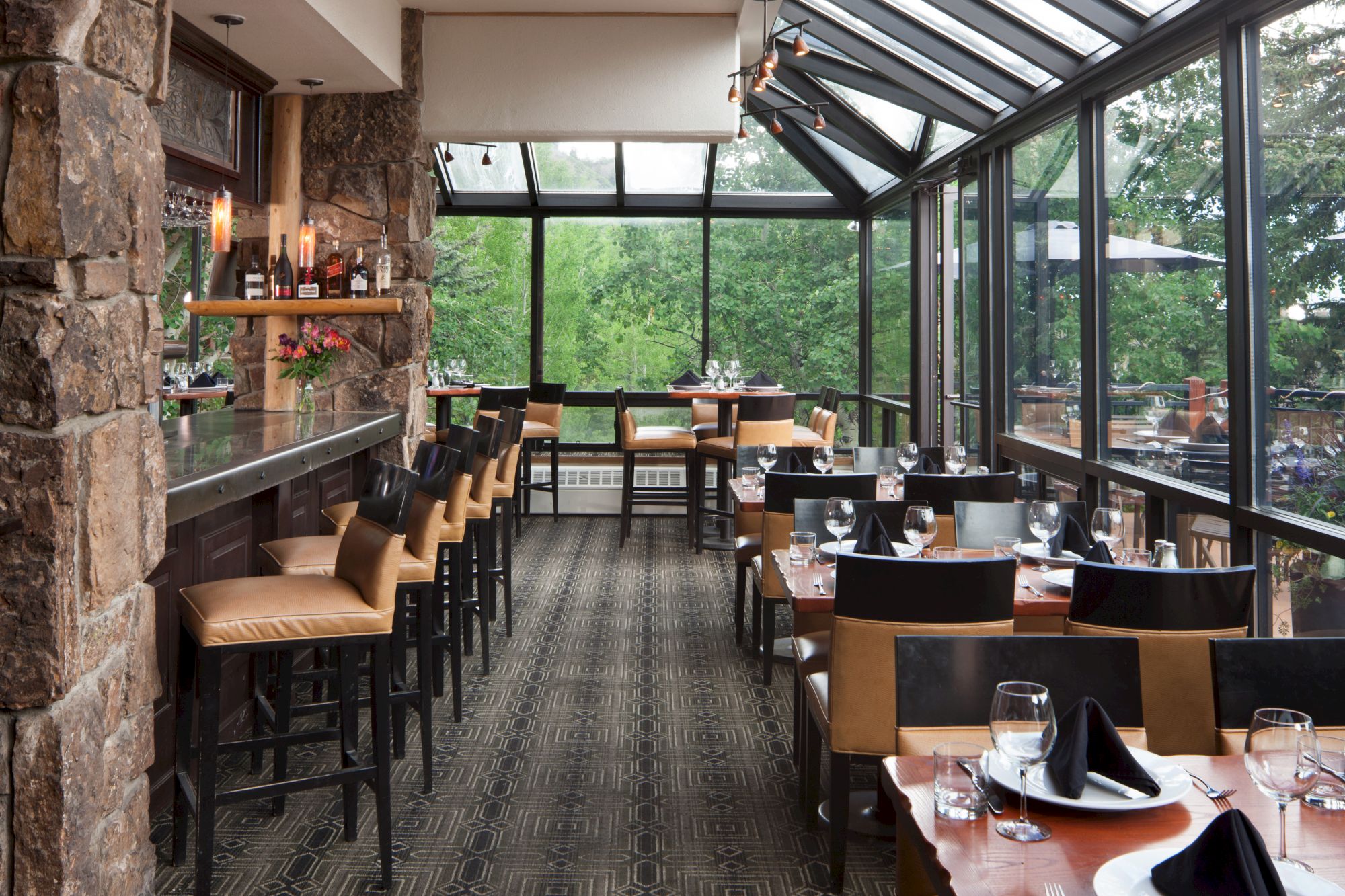 An elegant restaurant with a stone wall, bar seating, dining tables, large windows, and a view of trees. Table settings include napkins and glasses.