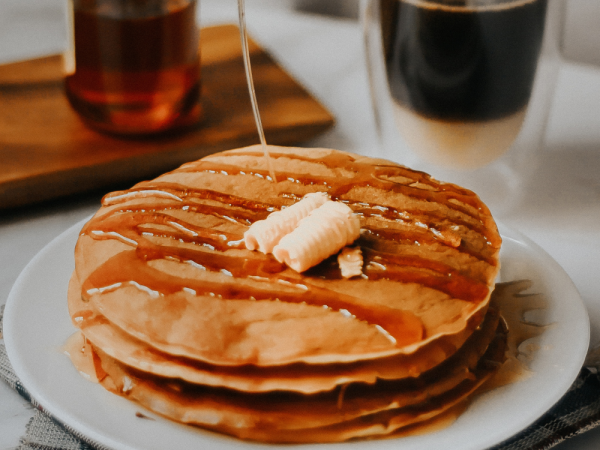 A stack of pancakes topped with butter and drizzled with syrup, alongside a cup of coffee and a bottle of syrup on a table.