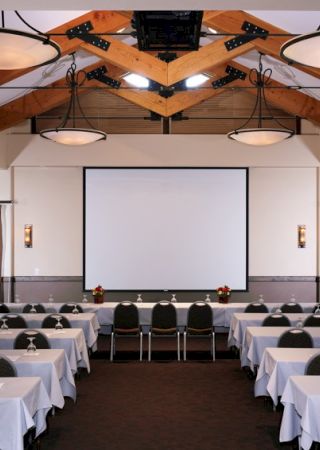 The image shows a conference room setup with rows of tables and chairs, facing a projector screen. The room features ceiling lights and wooden beams.