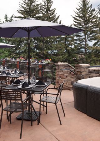 An outdoor patio with tables, chairs, umbrellas, and a couch seating area; surrounded by plants, trees, and scenic background.