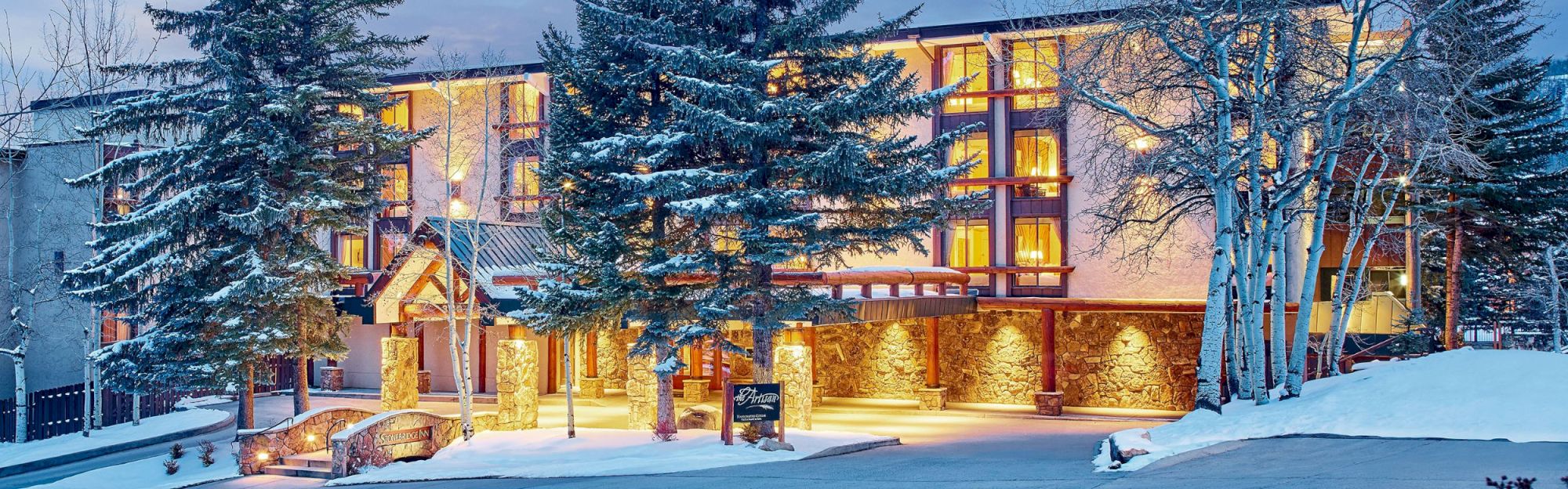 The image shows a beautifully lit building surrounded by snow and trees, likely a hotel or lodge, with a welcoming entrance and cozy ambiance.