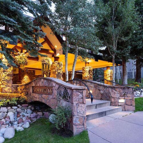 A warmly lit building entrance with stone pillars, surrounded by trees and landscaped greenery, under a twilight sky.