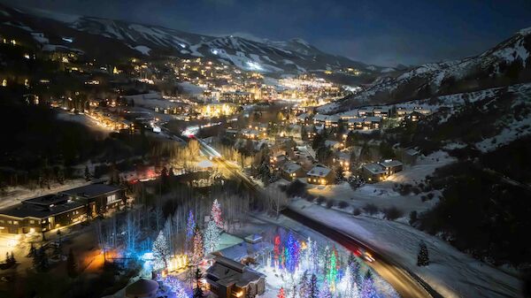 A snowy mountain town at night with colorful lights on a forested hill and winding streets, a festive glow against dark mountains.