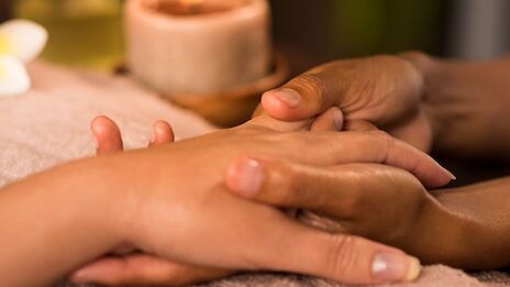 The image shows a hand massage being performed in a relaxing environment, with a lit candle in the background.