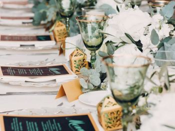A beautifully set table with green goblets, white flowers, decorative gold items, and menus placed at each setting, likely for a formal event or celebration.