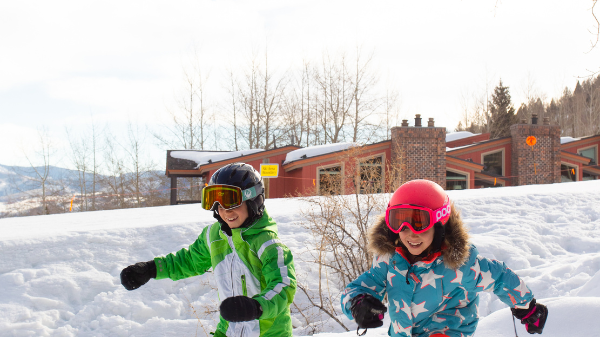 Two kids in colorful snow outfits are playing in the snow, while a ski gondola glides overhead in a winter setting.