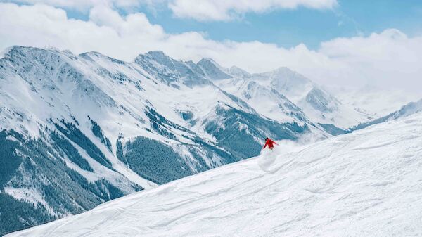 A lone skier in a red jacket carving down a snowy mountain slope with jagged peaks in the distance, bright skies above.