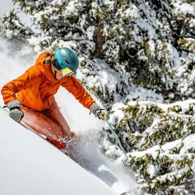 A skier in an orange jacket and teal helmet carves down a snowy slope, spraying powder as trees dusted with snow loom nearby.