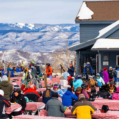 A crowded outdoor ski lodge scene with people seated at red-checked tables, kids in colorful gear, and mountains in the background, fun and festive.
