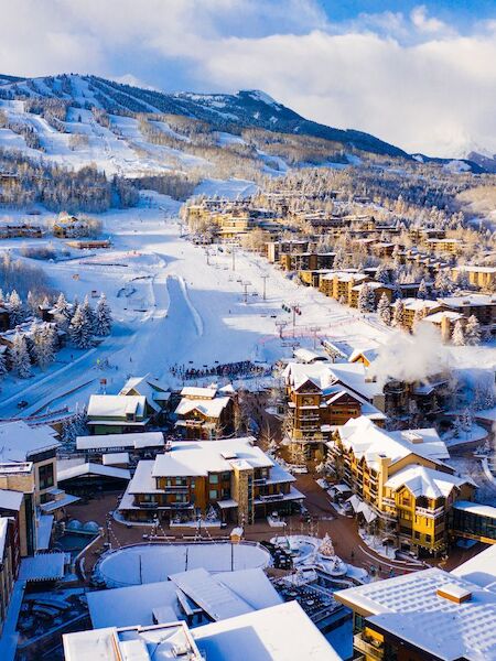 A snowy mountain village with chalets clustered along a winding road, framed by pine trees and pale blue sky, cozy and scenic.