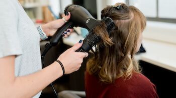 A person is blow-drying another person's hair using a round brush in a salon or similar setting.