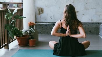 A person is practicing yoga on a mat indoors, seated with their hands in a prayer position behind their back, surrounded by plants.