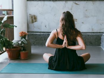A person is practicing yoga on a mat indoors, seated with their hands in a prayer position behind their back, surrounded by plants.