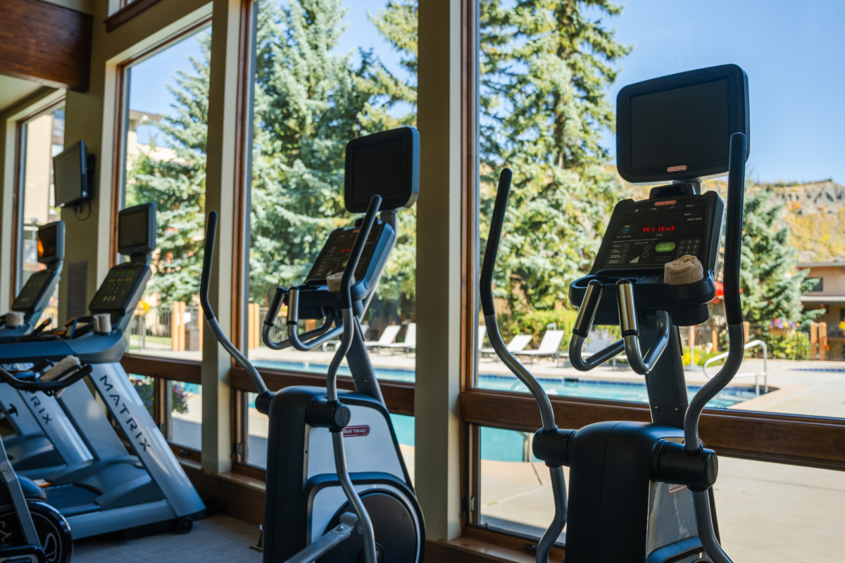 A gym with exercise equipment including stationary bikes and treadmills, located next to large windows showcasing trees and an outdoor pool area.