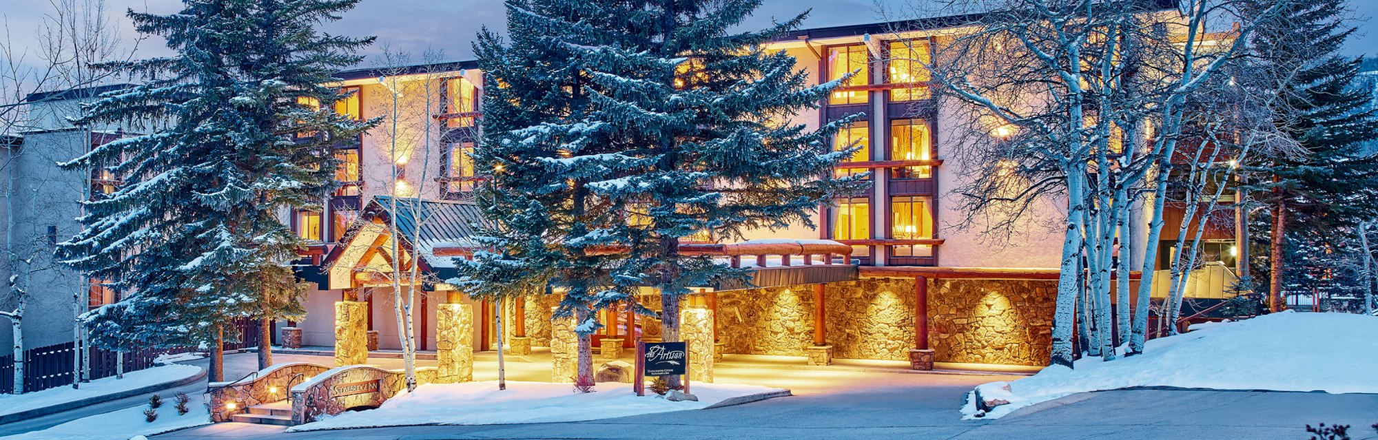 A warmly lit, multi-story lodge or hotel nestled in a snowy, forested area with large pine trees and an inviting entrance.