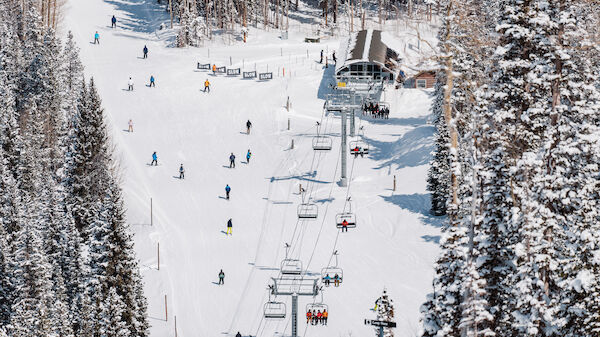 The image shows a snow-covered ski resort with skiers on the slopes and ski lifts in motion among snowy trees and a building in the background.